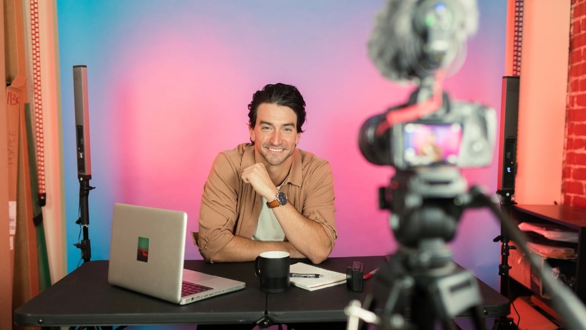Profissional sorridente gravando vídeo em estúdio com laptop e câmera, durante criação de conteúdo digital para marketing ou redes sociais.